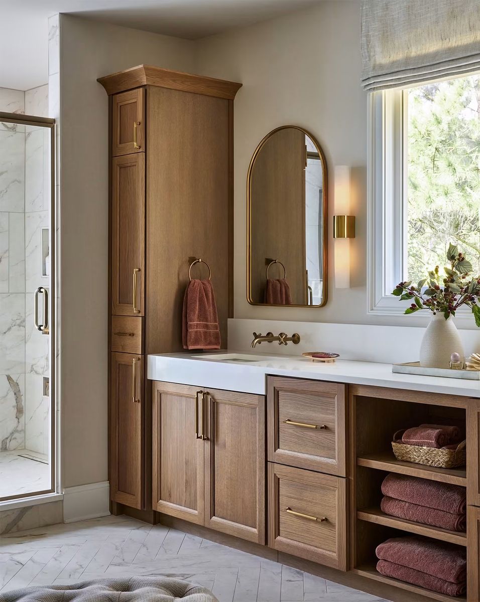 Oak bathroom vanity with a tall linen cabinet, open shelves, an arched brass mirror, wall-mounted brass faucet and a glass shower
